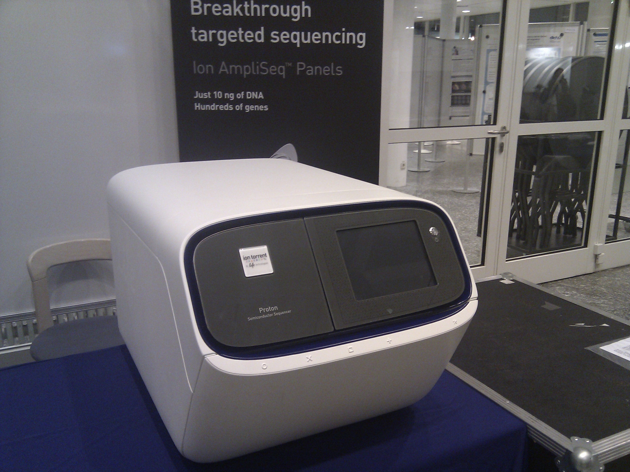 A modern DNA sequencing machine from Life Technologies sits on a blue tablecloth, with a display screen and Ion Proton labeling. Behind it, a banner reads Breakthrough targeted sequencing Ion AmpliSeq Panels.