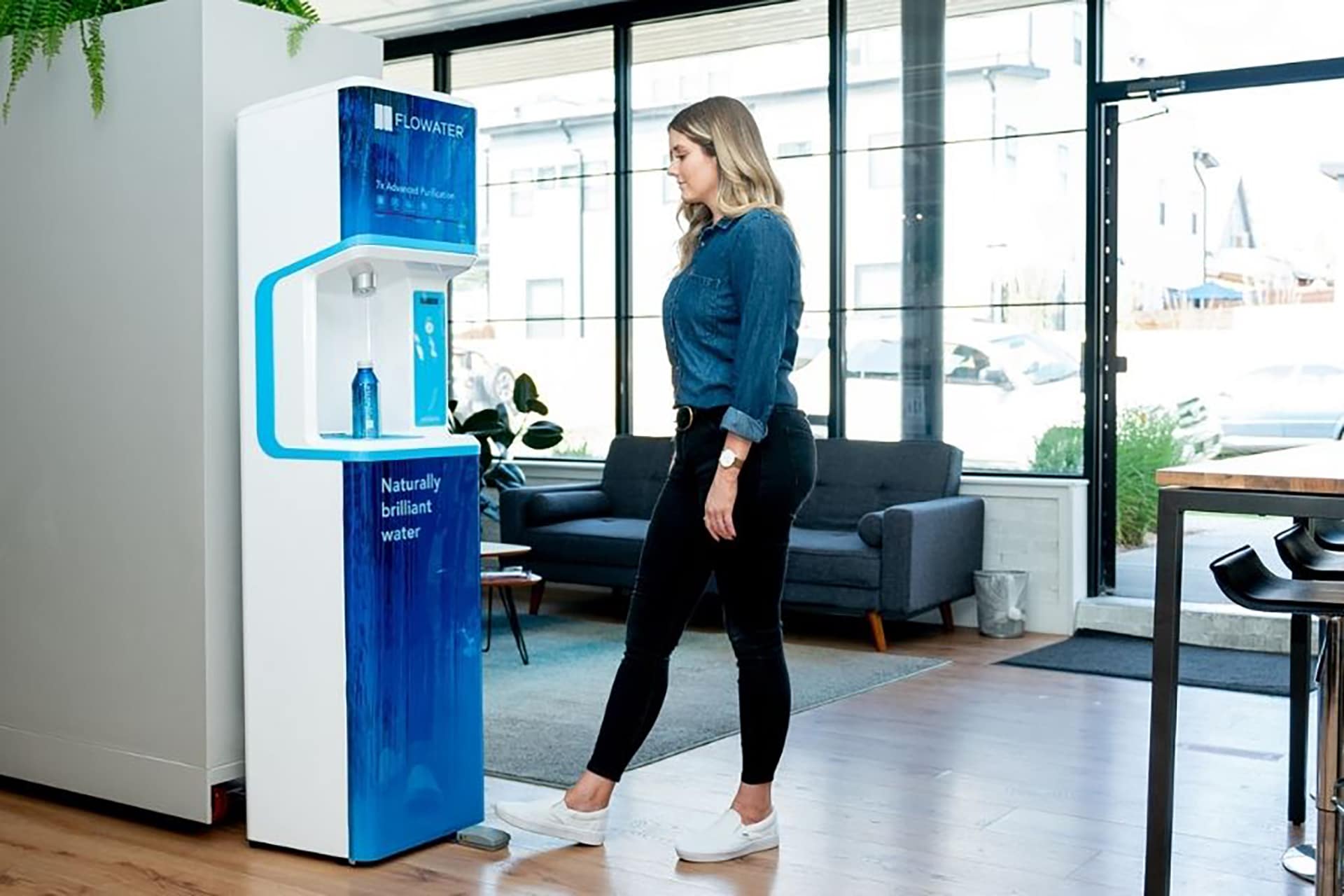A woman stands in front of a Flowater water dispenser, filling a reusable bottle in a modern, bright office with a sofa, chairs, and large windows in the background.