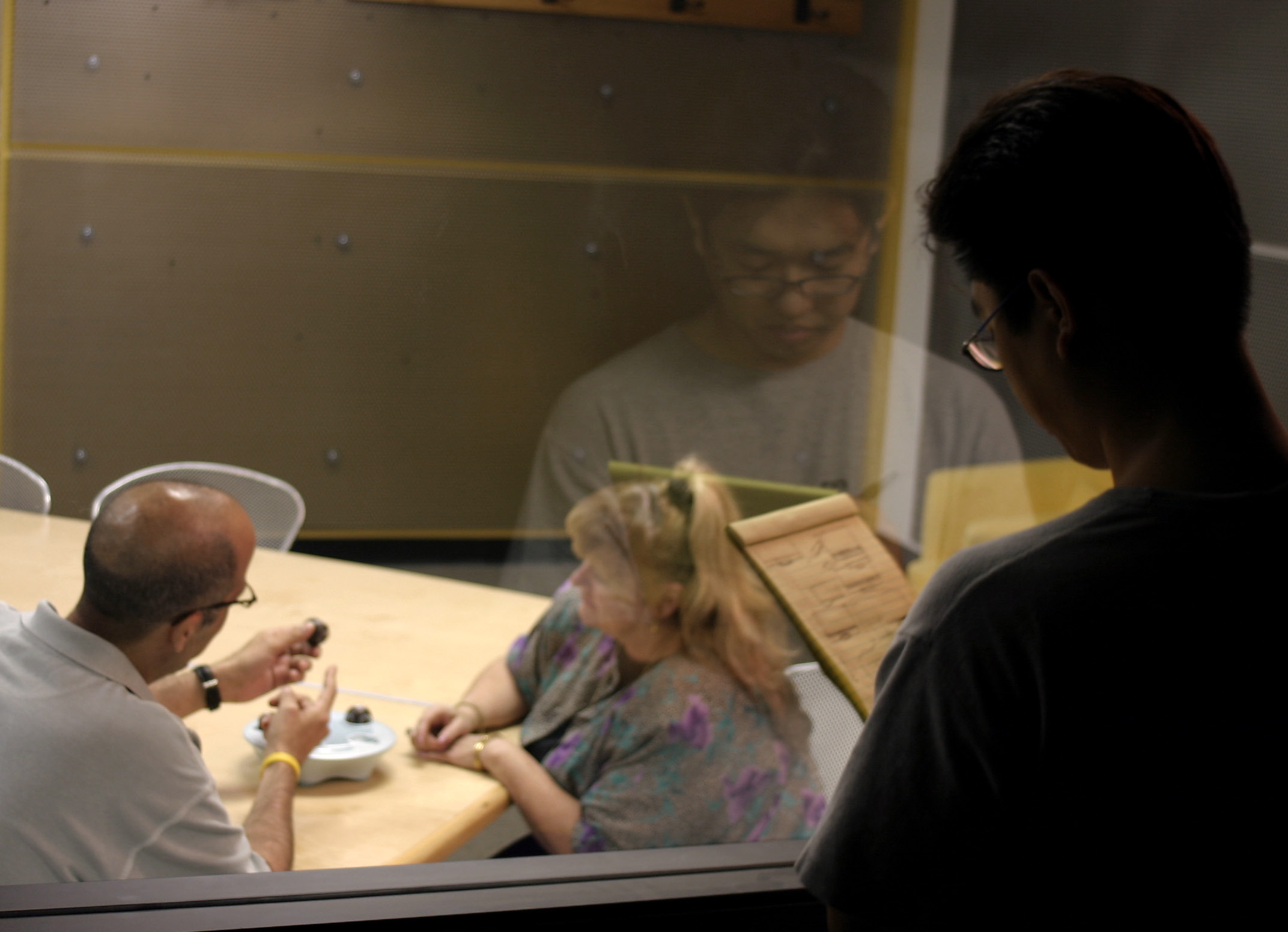 A man and a woman sit at a table discussing something, while another man observes and takes notes from behind a glass window. The scene appears to be in an observation or interview room.