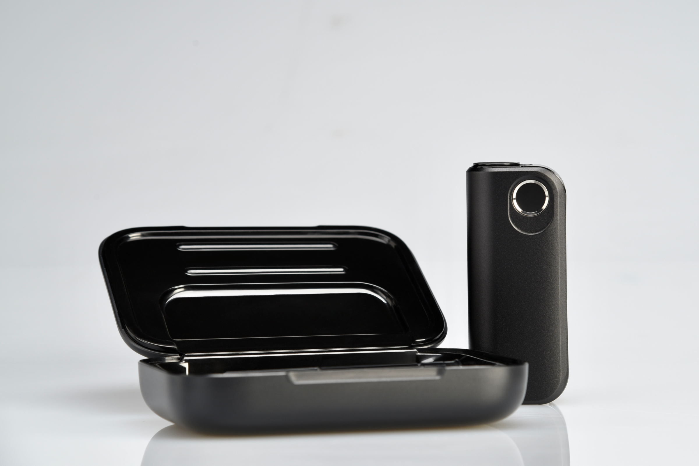 A sleek, black rectangular case is open and empty on a white surface, with a matching compact device standing upright next to it. Both items have a modern, minimalist design.