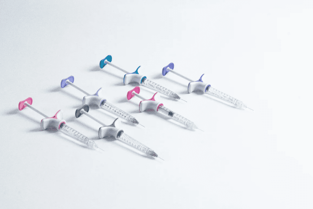 Six syringes with different colored plungers are arranged diagonally on a white surface. The colors of the plungers include purple, blue, and pink, and the syringes are spaced evenly apart.
