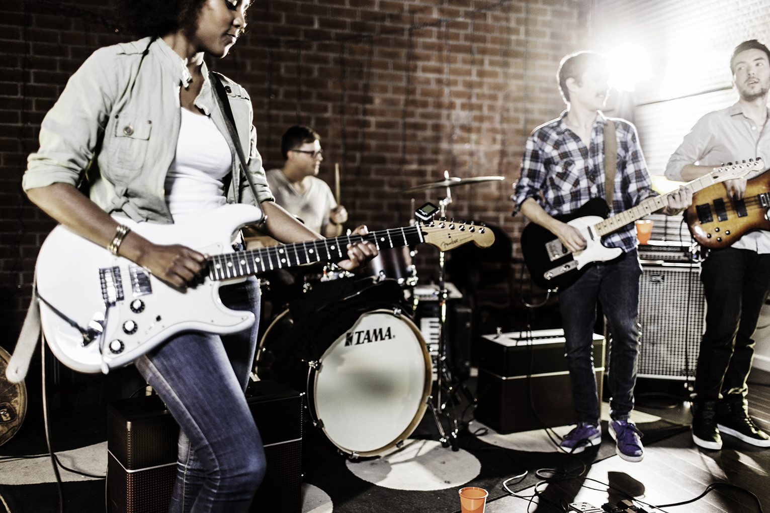 A band performs in a studio with brick walls; a woman plays electric guitar, a drummer is at a drum set, and two men play electric and bass guitars under bright stage lights.