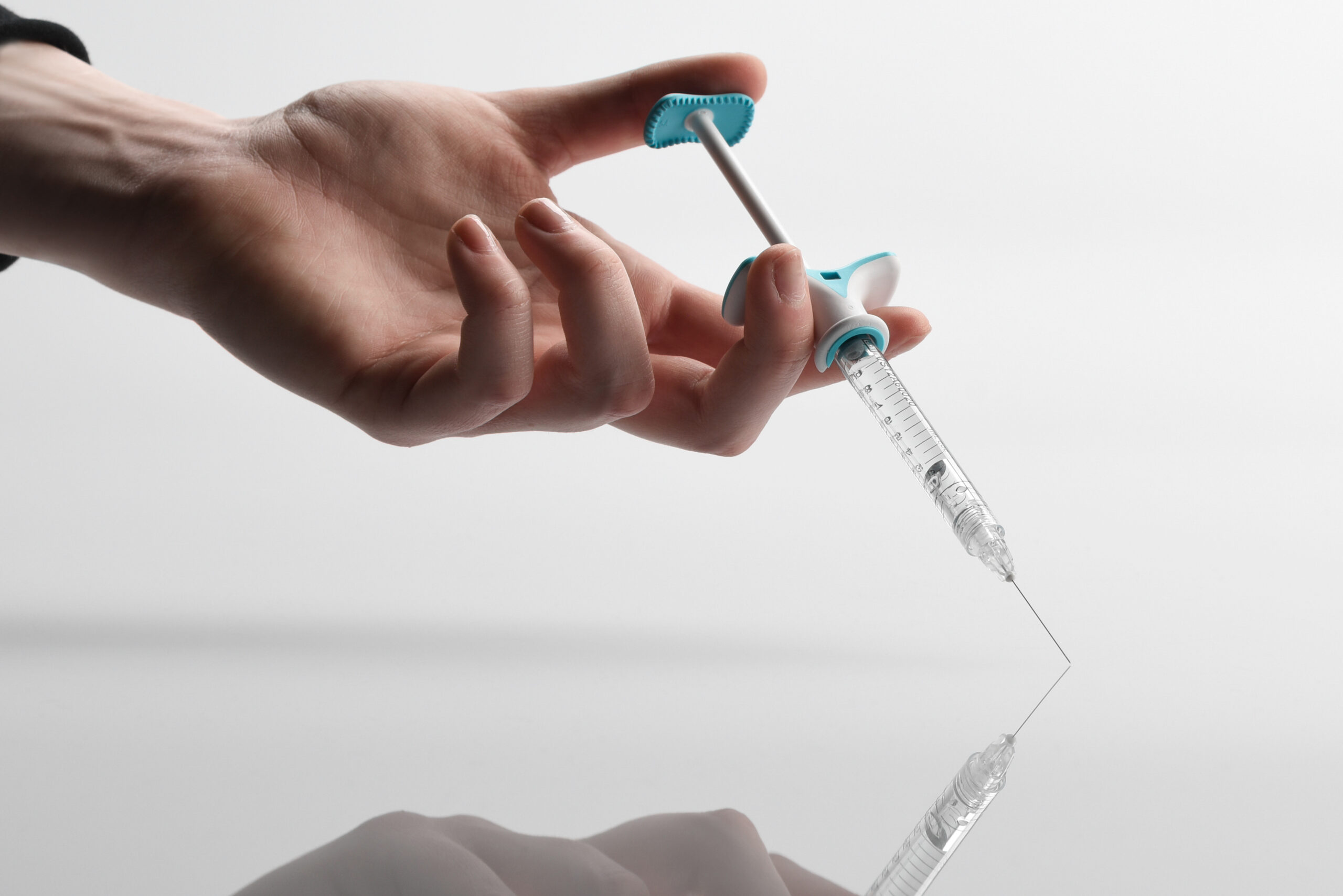 A hand holds a syringe with a blue plunger, pressing it down as the needle touches a reflective surface against a white background.
