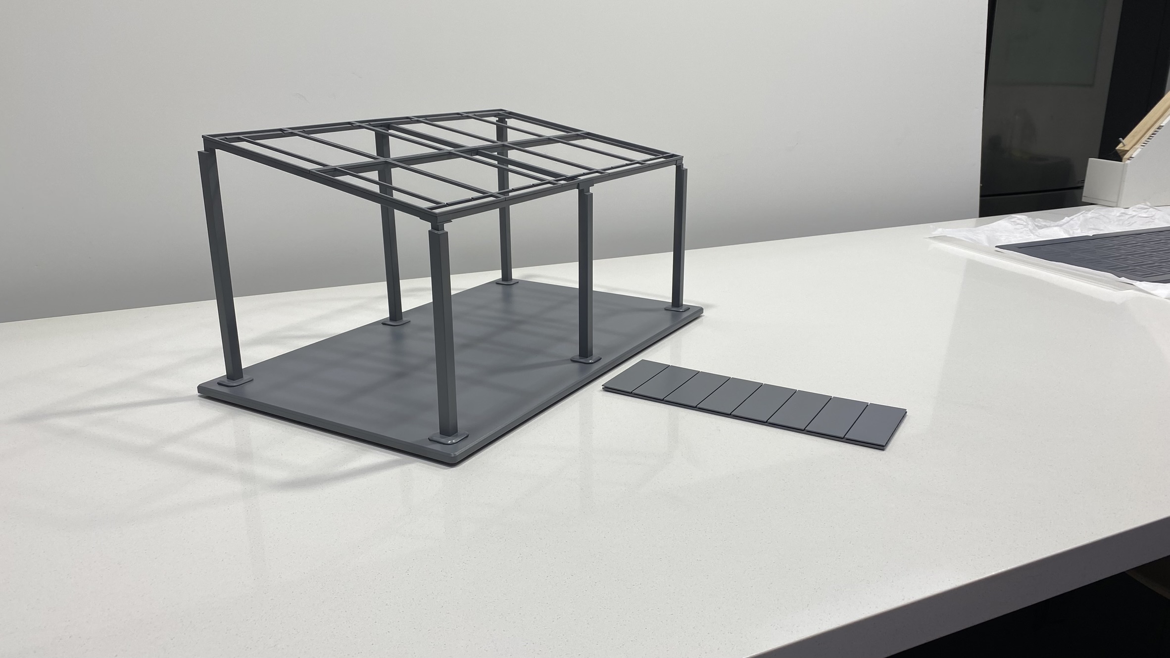 A small, gray, rectangular model frame with six vertical posts and a grid roof structure sits on a white surface. A separate flat panel with grooves lies next to the model.