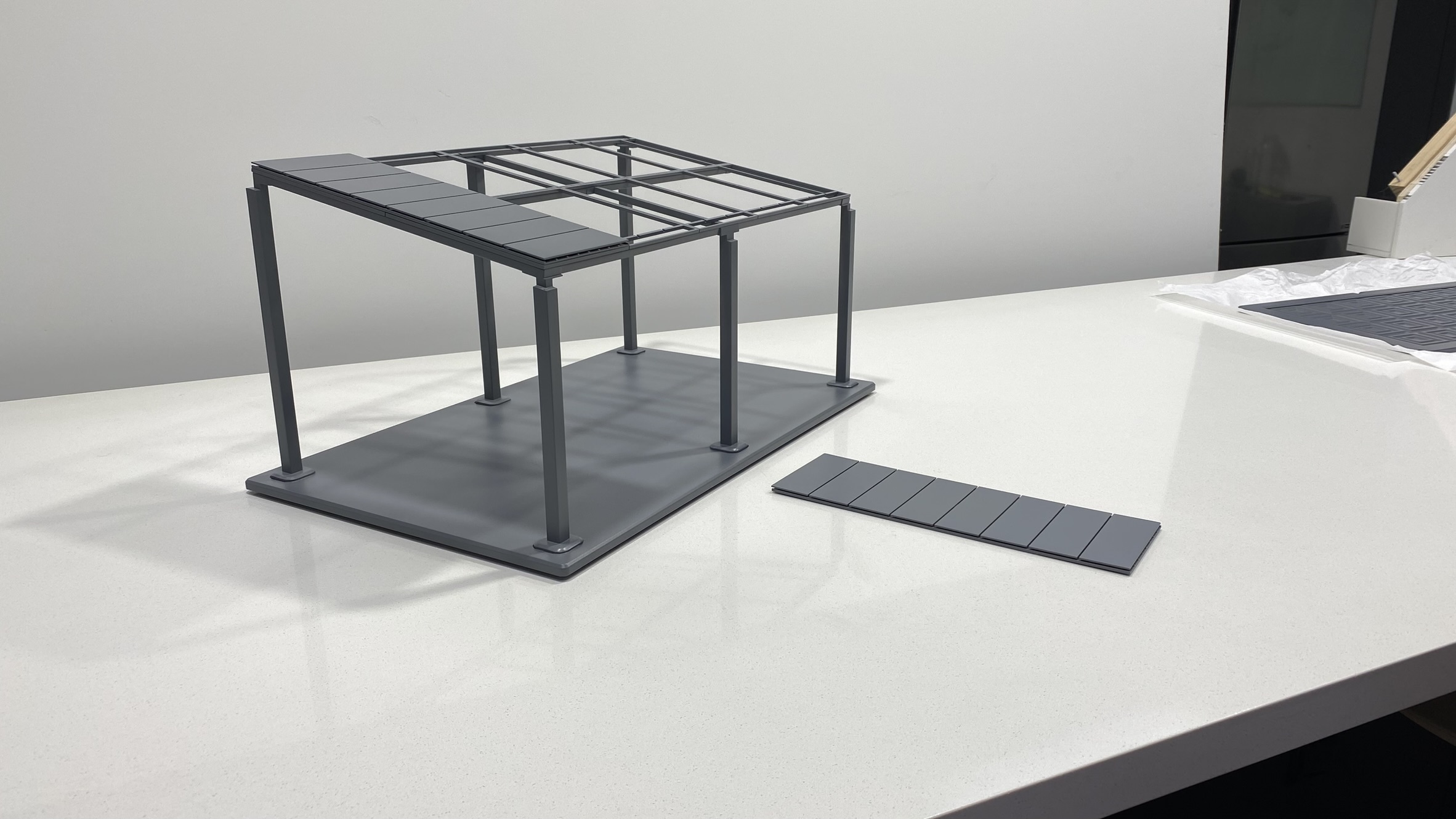 A small gray model structure with a rectangular base and several vertical and horizontal beams stands on a white surface. A detached roof panel lies next to the model.
