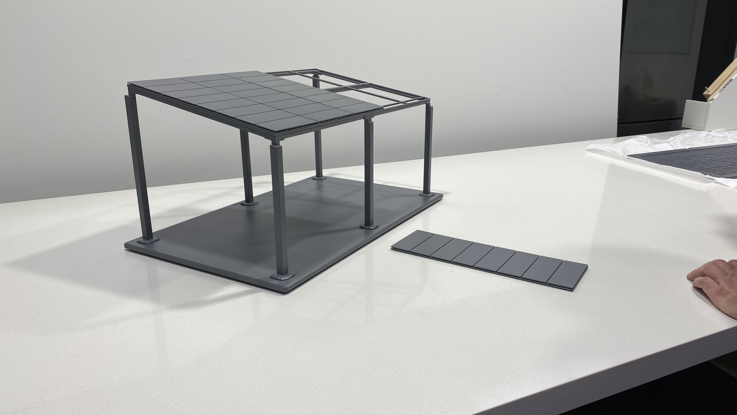 A scale model structure with a rectangular base and four vertical posts supports a partially constructed roof. A separate panel lies flat on the white surface next to the model, and a hand is visible at the right edge of the image.
