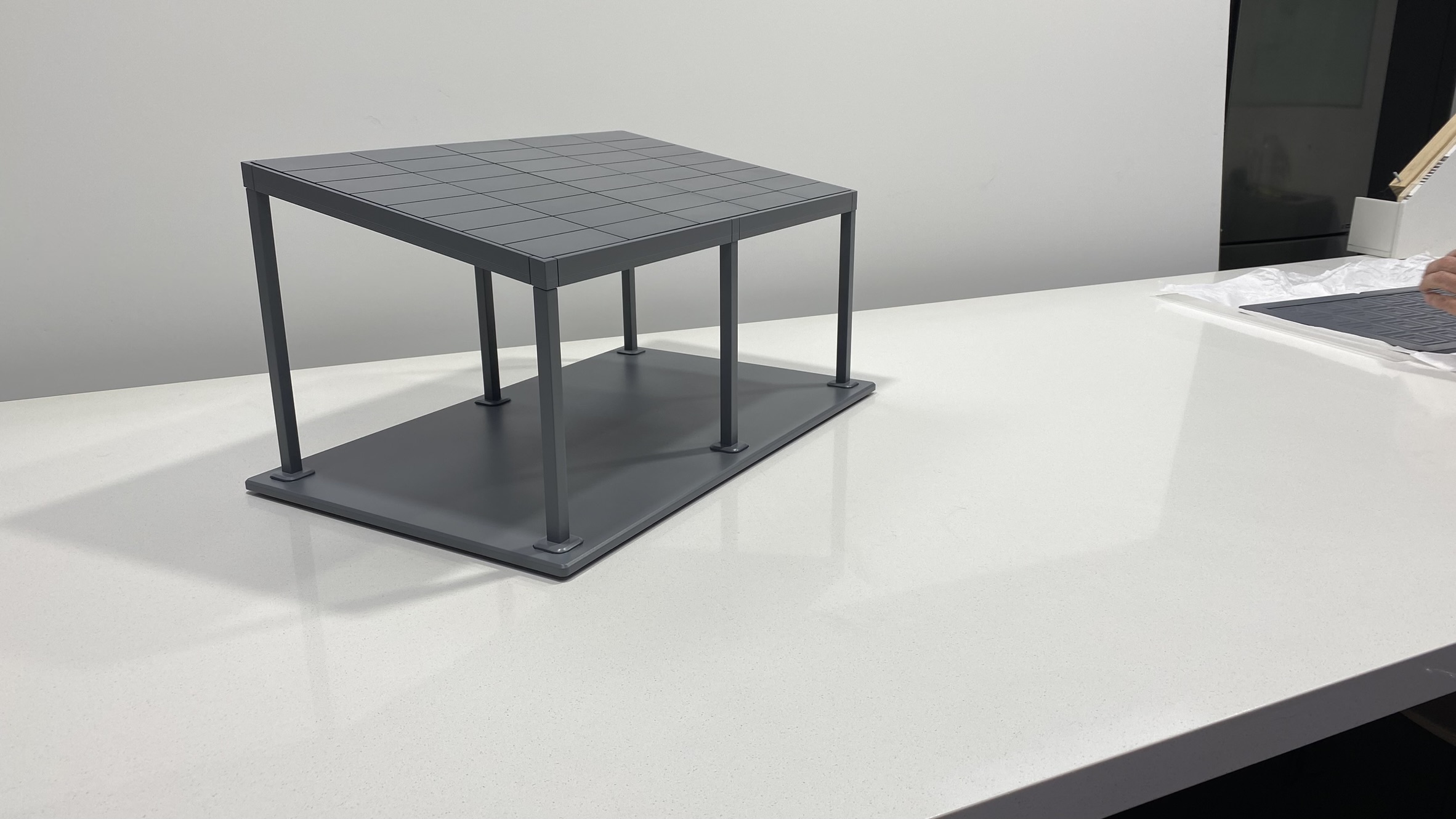 A gray plastic model of a rectangular table with four legs stands on a white countertop. The tabletop has a grid pattern, and some materials are partially visible in the background.