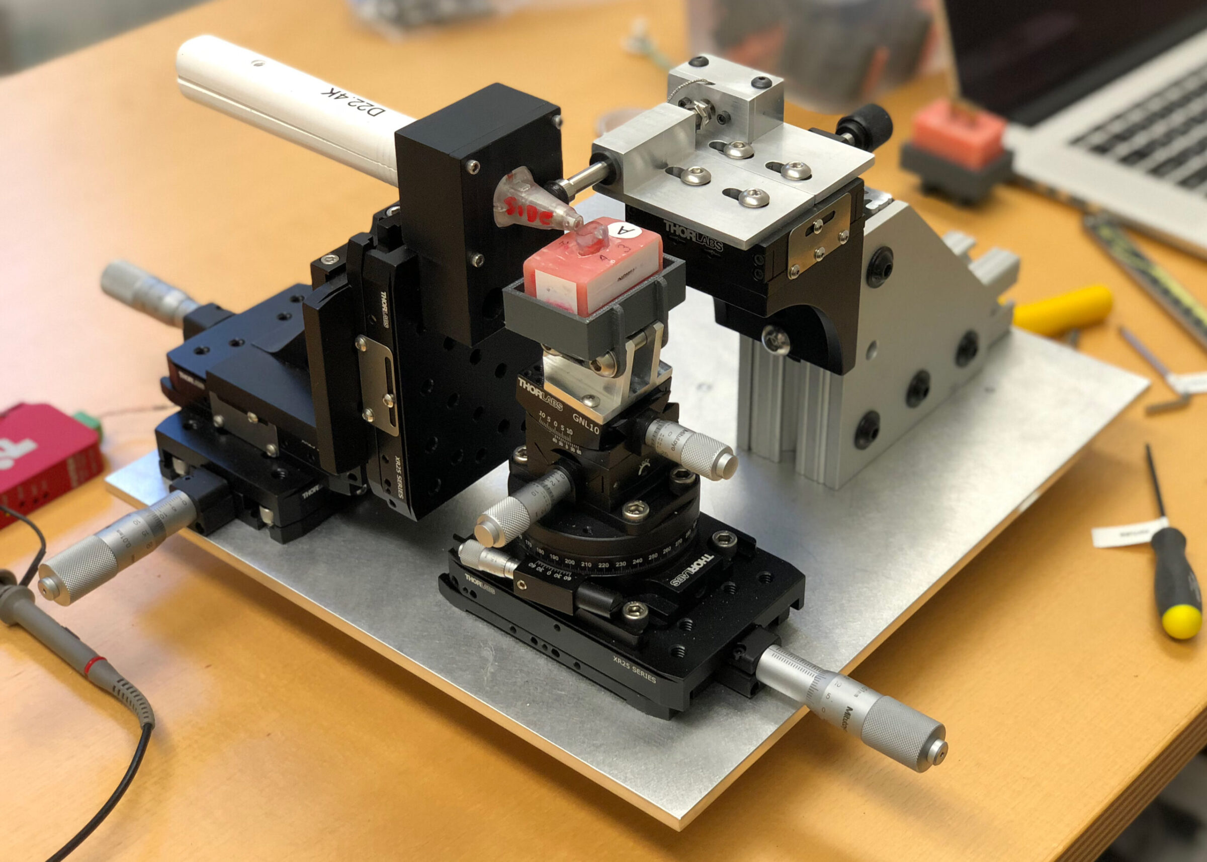 A close-up of a precision scientific instrument with micrometer controls and mechanical components, set on a metal baseplate on a wooden desk, with tools, a laptop, and Perimetrics analytics running in the background.