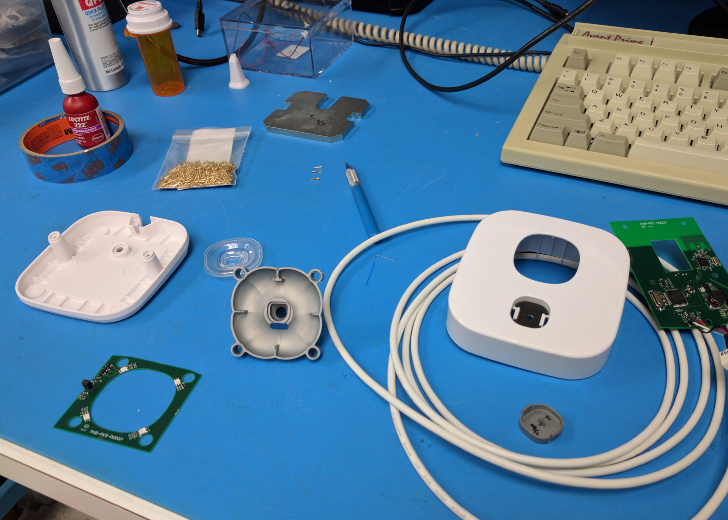 An electronics workbench with scattered components: a disassembled white plastic device, circuit boards, a coiled cable, a craft knife, screws, and tools on a blue surface next to a beige keyboard—perfect for Perimetrics or InnerViewAI projects.