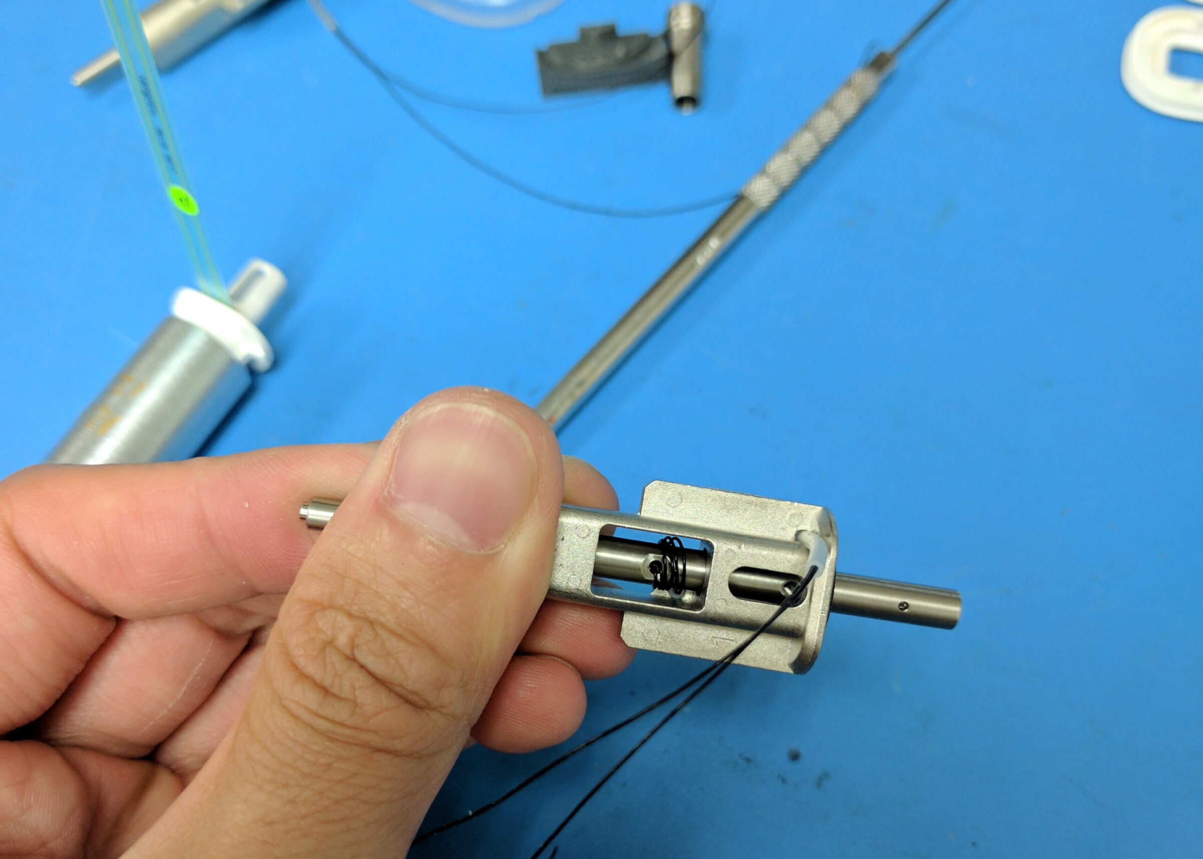 A hand holds a small metal mechanical component with a spring and rod, against a blue background with various metal tools and Perimetrics parts scattered nearby.
