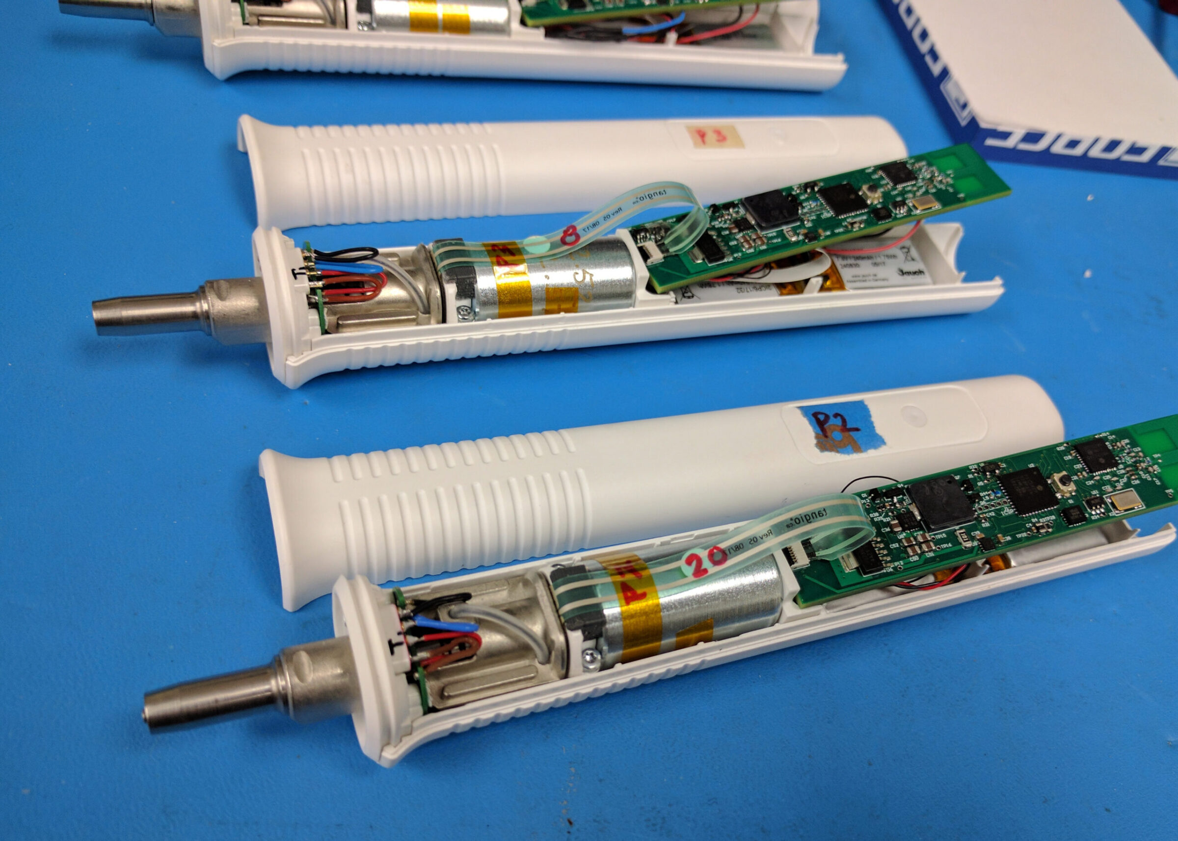 Three partially assembled electronic handheld devices with white plastic cases—such as those used in InnerViewAI or Perimetrics—are lying on a blue surface, exposing internal circuit boards, wiring, and cylindrical batteries.