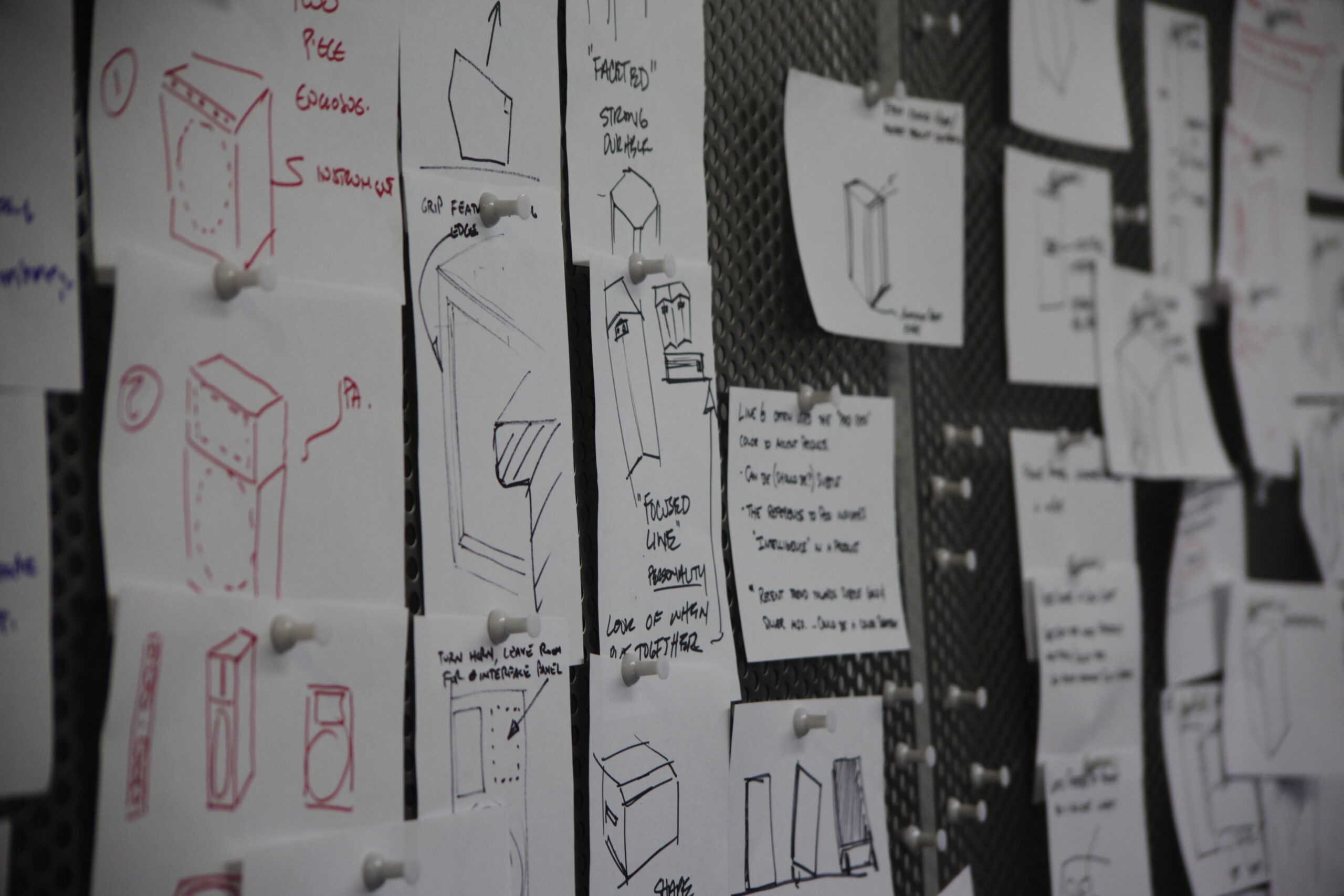 A bulletin board covered with sheets of paper displaying various hand-drawn sketches and notes, mainly of box-like structures, attached with push pins.