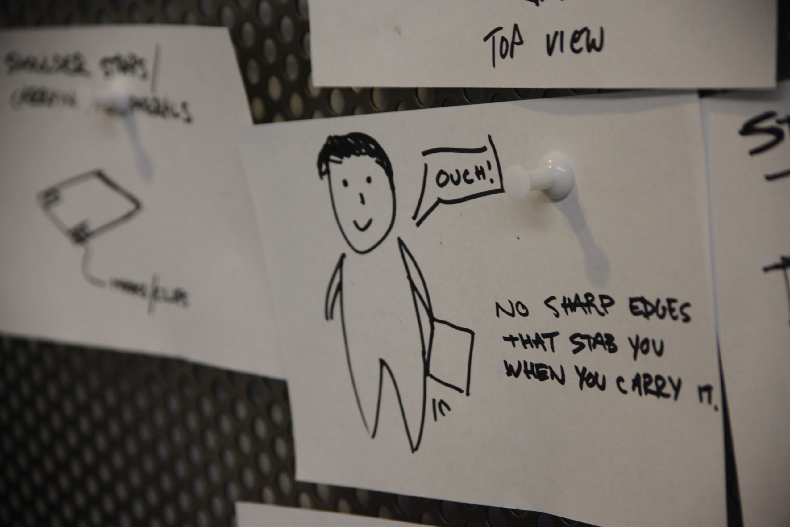 A hand-drawn sketch of a person holding a bag, saying Ouch! The note beside reads, No sharp edges that stab you when you carry it, and is pinned to a board among other sketches.