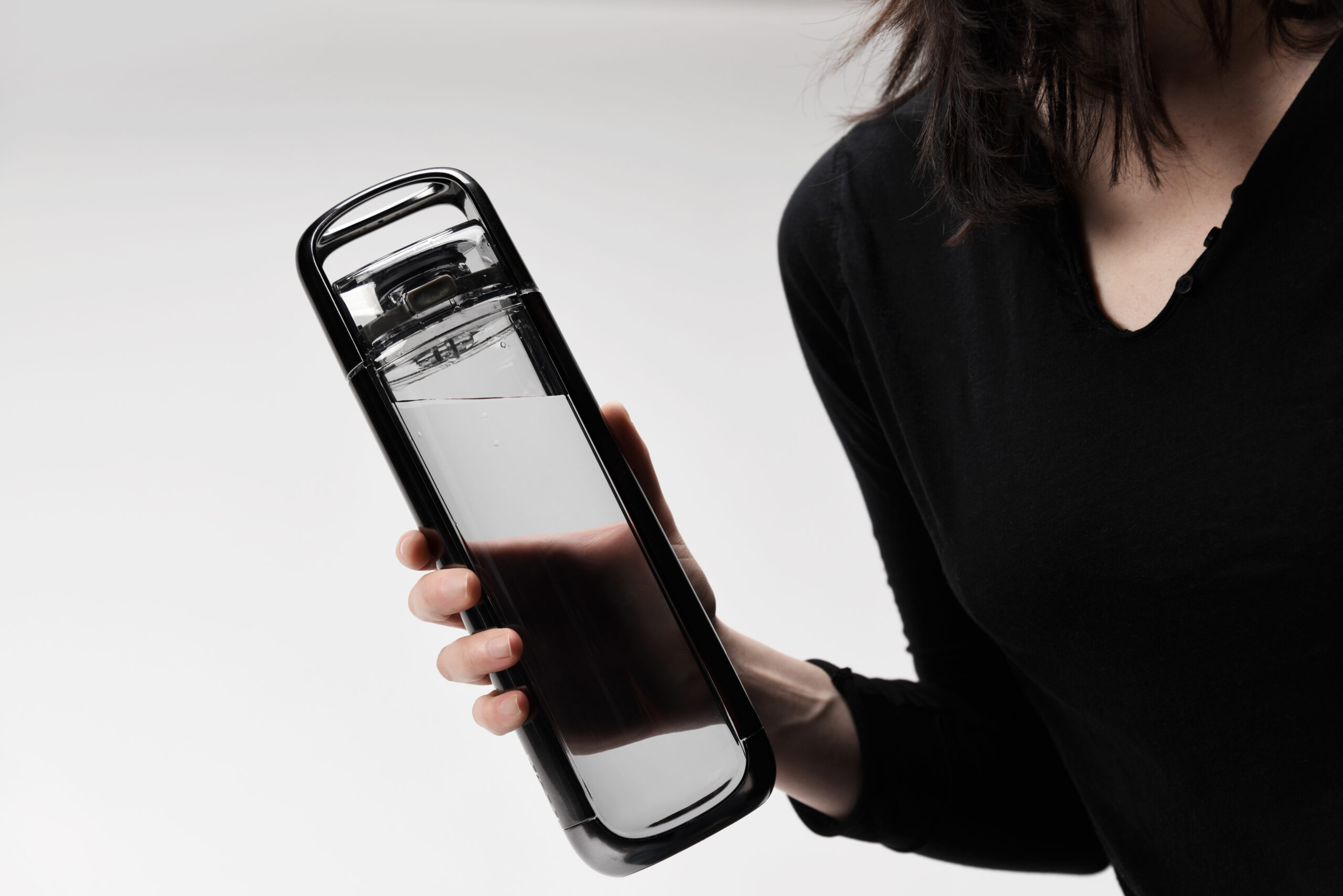 A person in a black long-sleeve top holds a clear water bottle with black accents, filled with a dark liquid, against a plain white background.