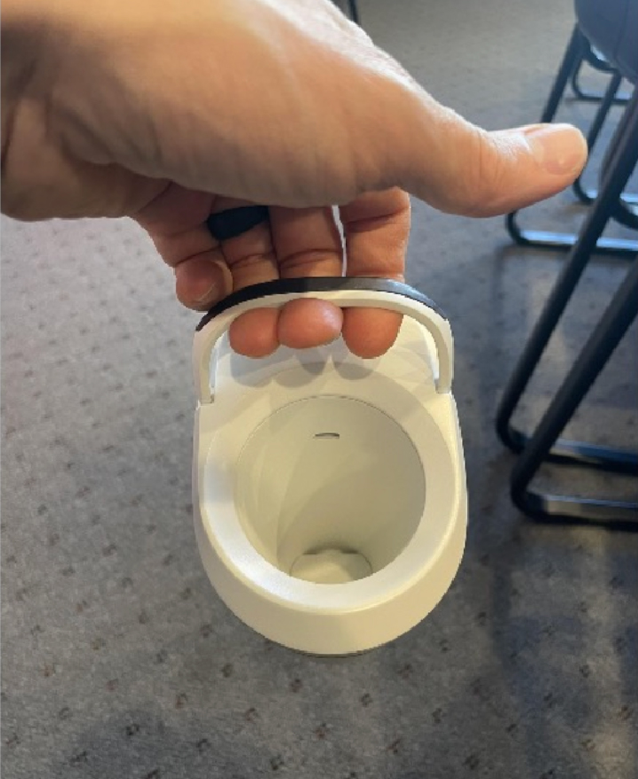 A hand holding a small, white, toilet-shaped object by its lid handle, above a carpeted floor with chairs in the background.