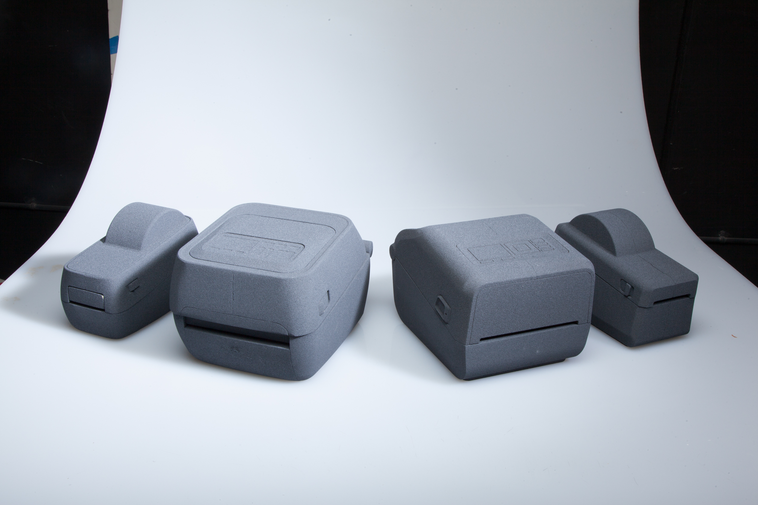 Four gray, rectangular devices with rounded edges are arranged in a row on a white curved surface. The devices appear identical in pairs, each pair differing in size and shape.