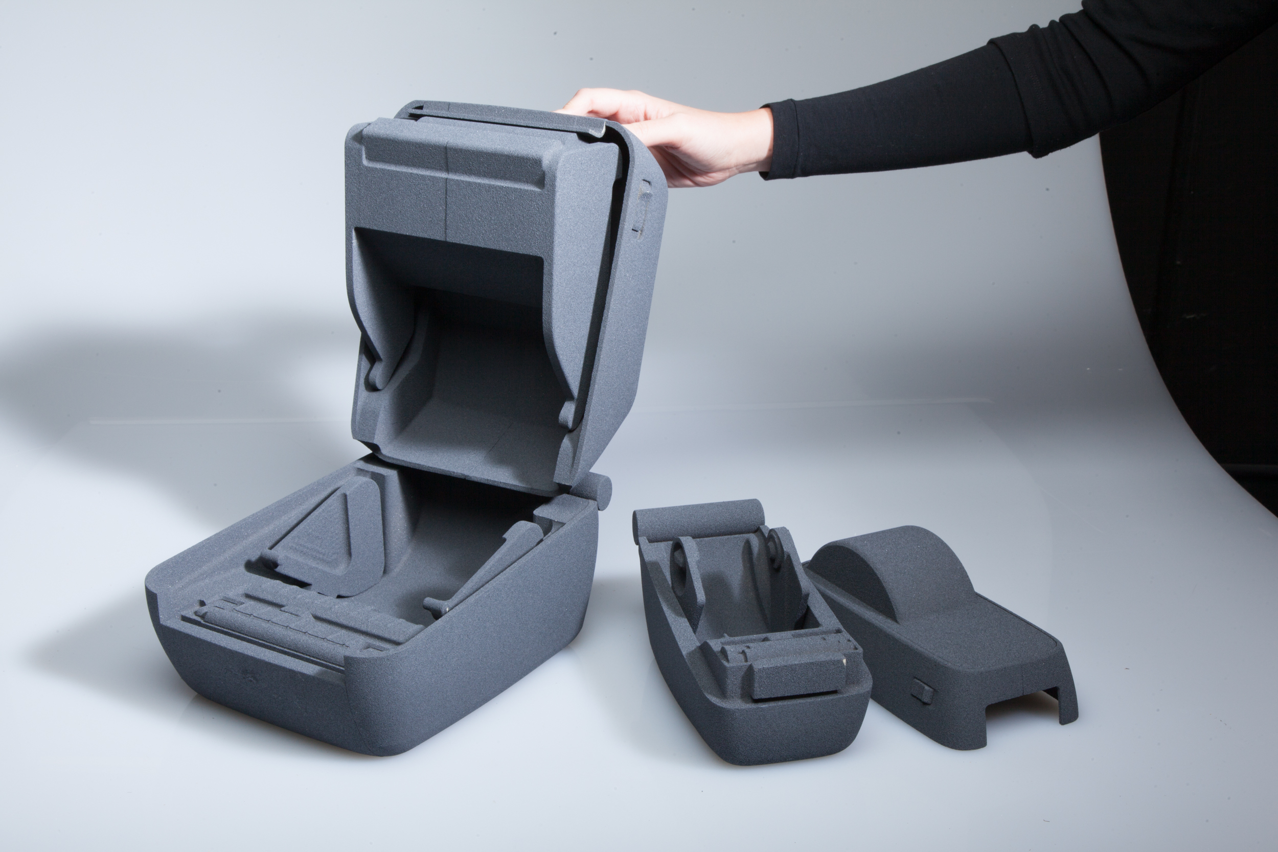 A person’s hand opens a large, gray, foam storage case next to two smaller, similar foam inserts on a white surface. The objects appear to be protective packaging or custom inserts.