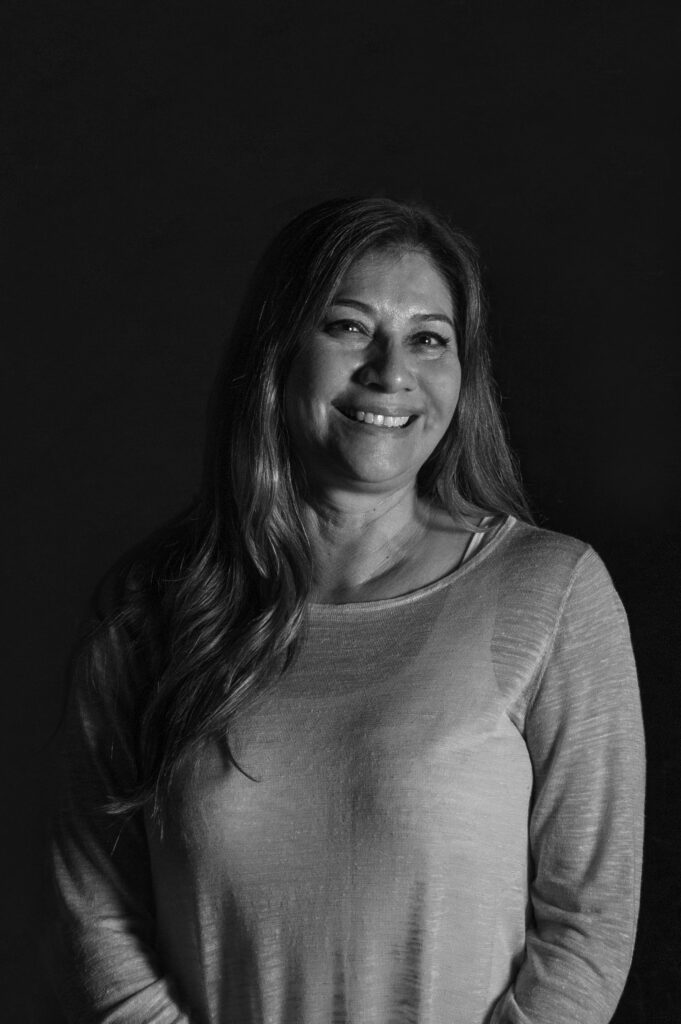 Black and white photo of a woman with long hair, smiling and facing the camera. She is wearing a light, long-sleeved top and stands against a dark, plain background.
