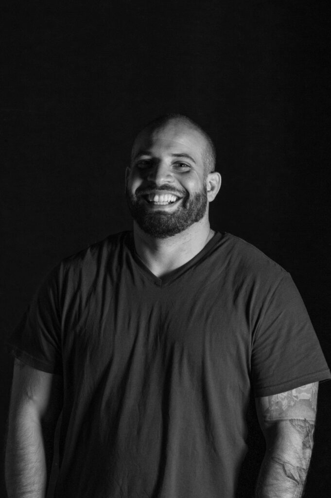 A man with a beard and short hair smiles broadly, wearing a plain t-shirt. His arms, some with visible tattoos, hang relaxed at his sides. The background is dark, and the image is in black and white.