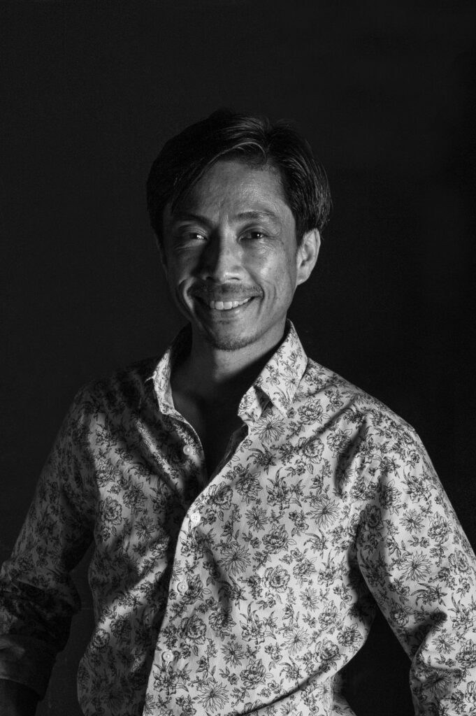 Black and white portrait of a smiling person wearing a floral-patterned shirt, standing against a dark background with light highlighting one side of their face.
