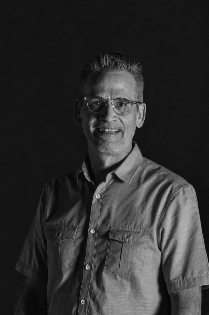 Black and white portrait of a middle-aged man with short hair and glasses, smiling and wearing a short-sleeve button-up shirt with chest pockets, standing against a dark background.