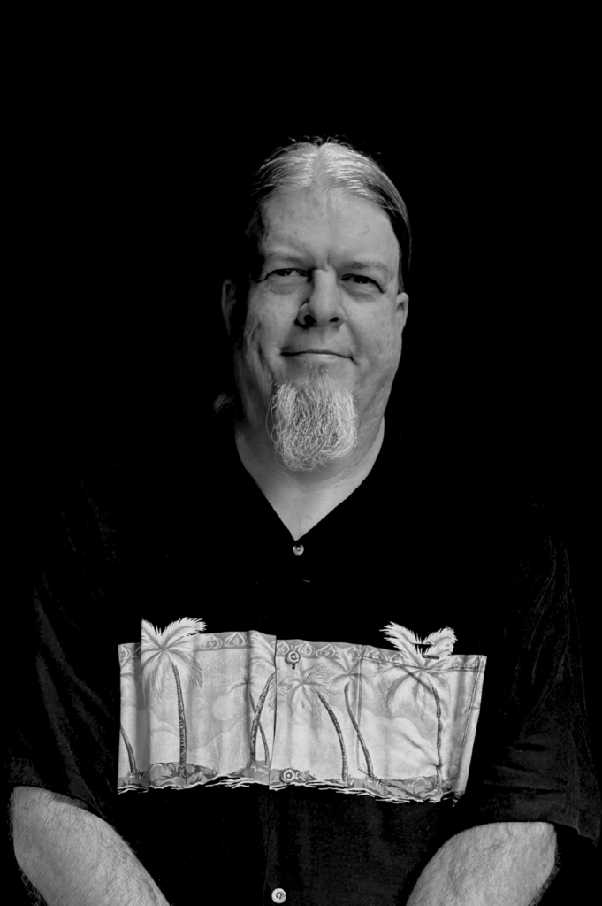 A man with long hair and a goatee stands against a black background, wearing a black shirt with a palm tree design across the chest. The image is in black and white.