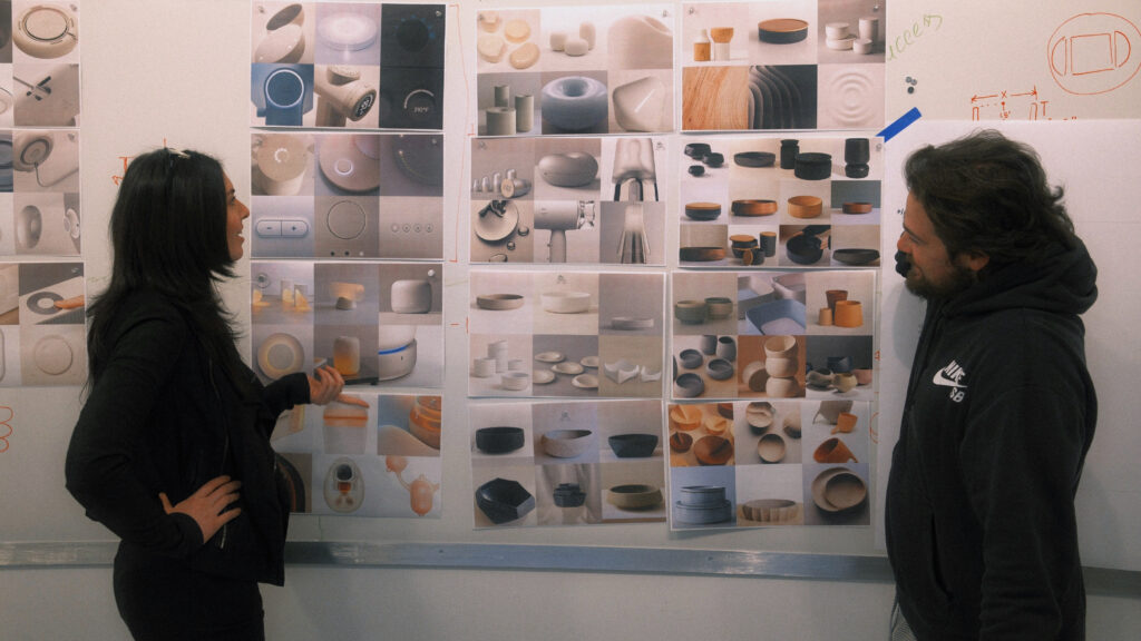 Two people stand in front of a wall covered with photos of ceramic objects, discussing the designs. One points to the images while the other looks at them thoughtfully.