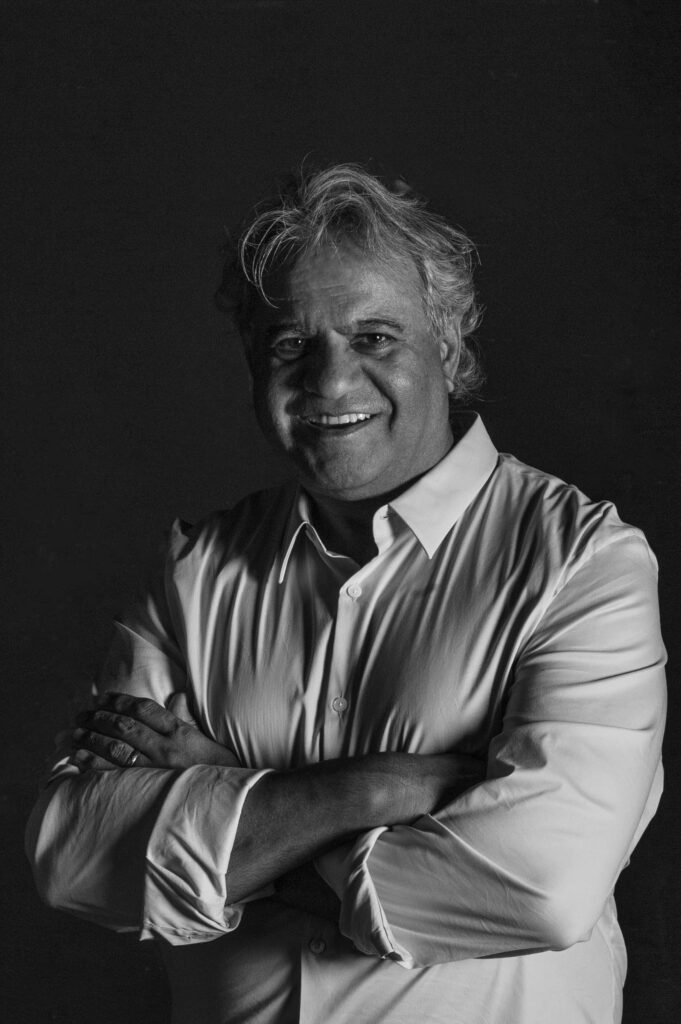 A man with gray hair wearing a light-colored button-up shirt stands with arms crossed, smiling, against a dark background in a black-and-white portrait.