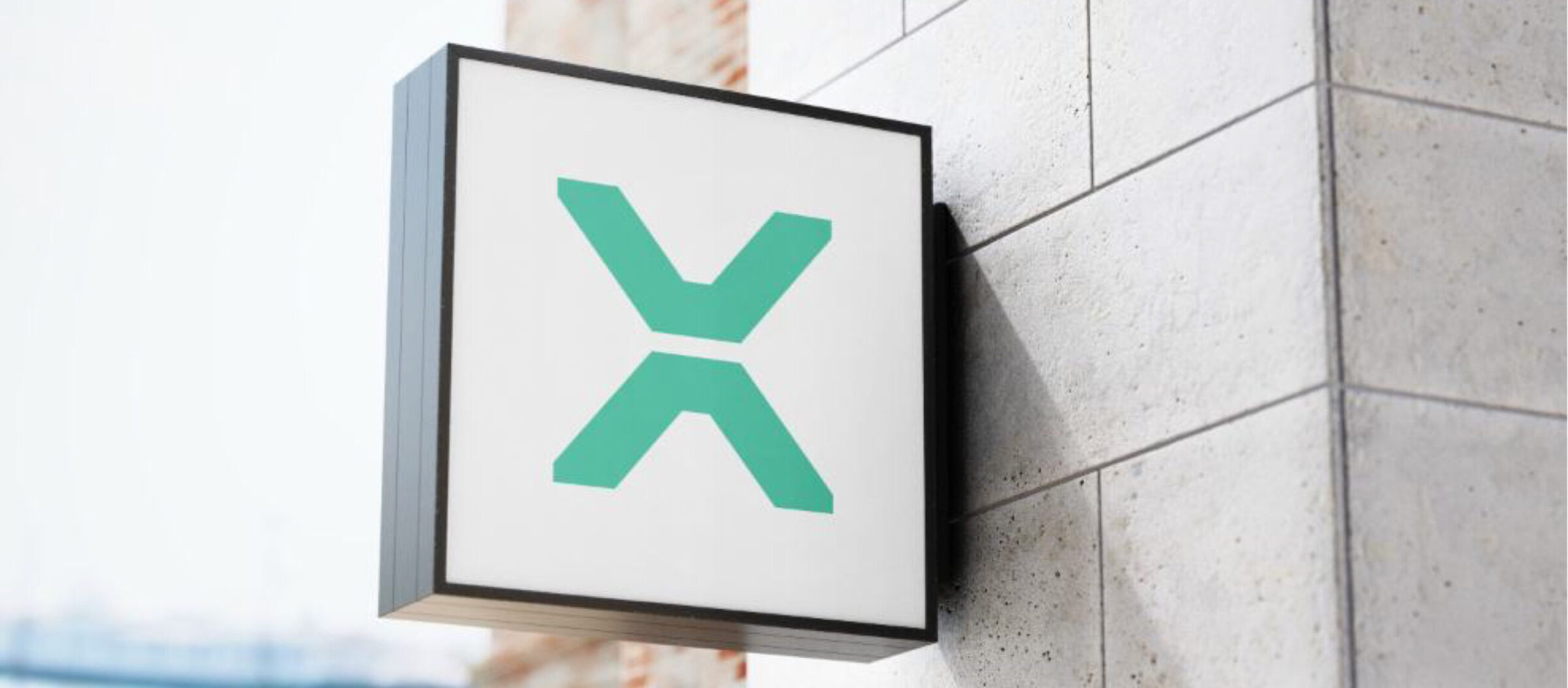 A wall-mounted square sign displays a green stylized X on a white background, attached to a light gray tiled building exterior.