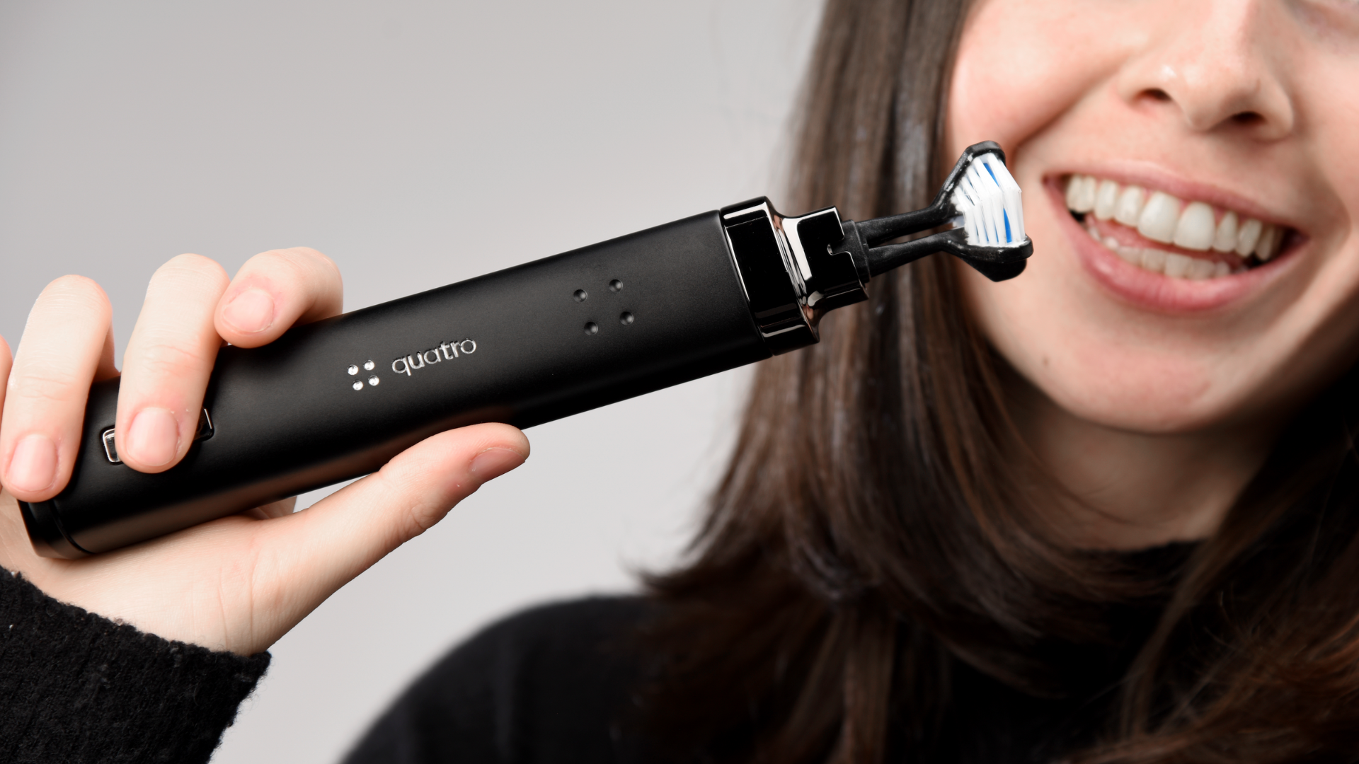 A smiling woman holds a black electric toothbrush with a triangular brush head near her mouth, preparing to brush her teeth.