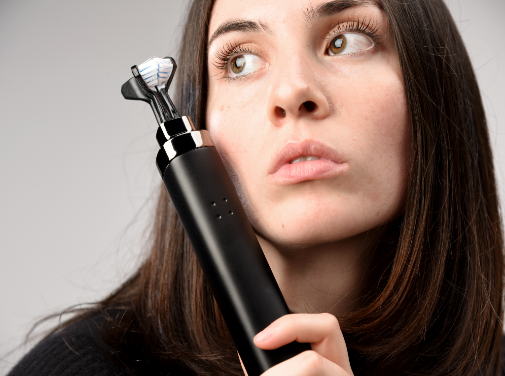 A woman with long brown hair holds an electric toothbrush near her face, looking off to the side with a thoughtful expression. The background is plain and neutral.