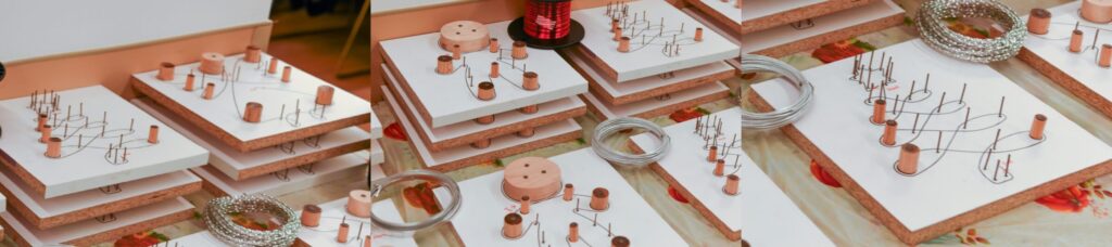 Stacks of white boards with wooden pegs and coils of metal wire are arranged on a table, ready for a Holiday Ornament Workshop. The background shows more supplies and a patterned tablecloth, adding to the creative momentum.