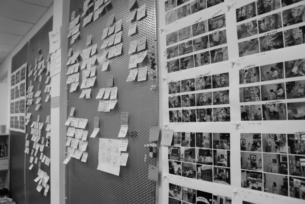 Black-and-white photo of walls covered in sticky notes, sketches, and printed photo sequences, suggesting a creative planning or brainstorming session in a workspace.