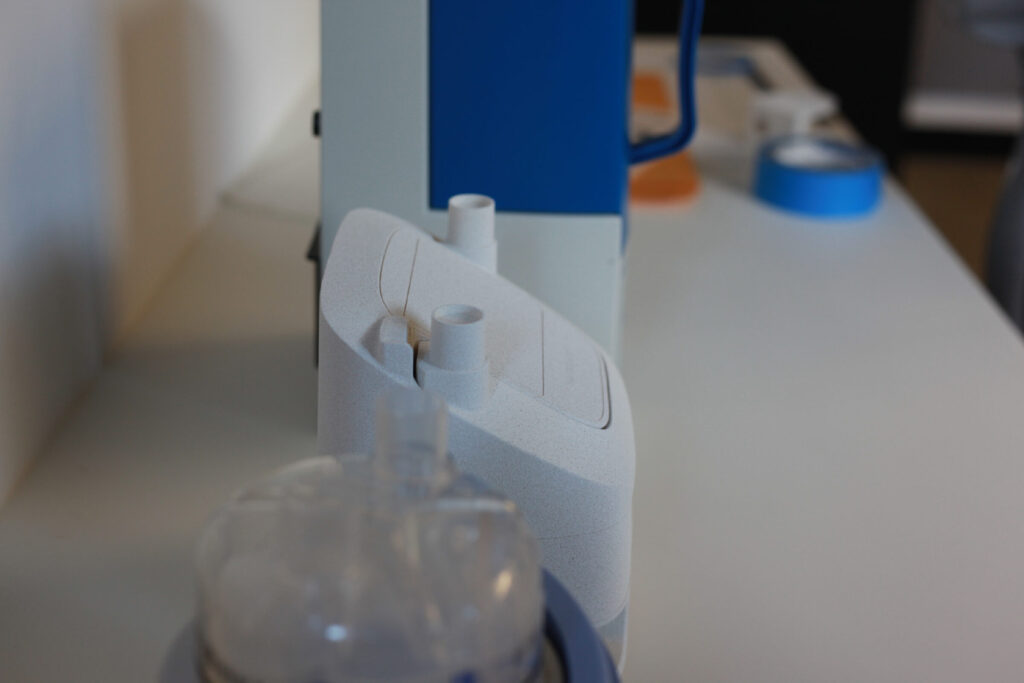 Close-up of a white medical device with cylindrical connectors on top, placed on a white surface. In the foreground, there is part of a transparent plastic component, possibly part of the same equipment.